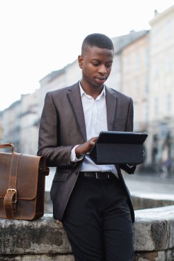 Takım elbiseli ve pantolonlu genç bir Afrikalı Amerikalı dışarıda duruyor. Elinde deri bir çanta, tablet cihazına odaklanmış.