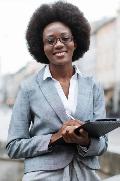 Afro saç stili ve gözlüklü profesyonel bir siyahi kadın, elinde bir pano ve kalem tutarken gülümsüyor ve iş ya da akademik bir ortam öneriyor.
