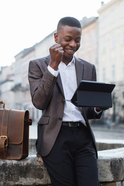 Takım elbiseli gülümseyen genç bir adam, bir şehir caddesinde evrak çantasını tutarken heyecanla tabletine bakar.
