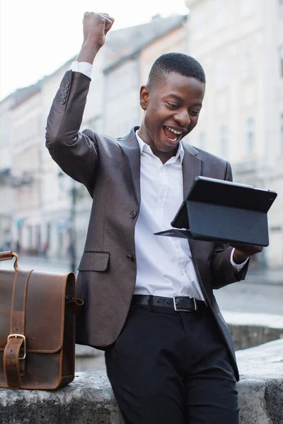 Takım elbiseli genç siyahi bir adam şehirdeki bir caddede tablet bilgisayara bakarken başarıyı kutluyor.