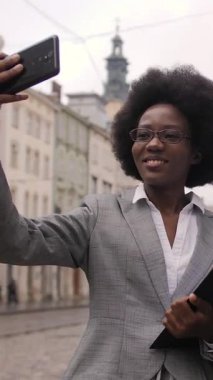 Başarılı afro-amerikan iş adamları şehir caddesinde yan yana durur ve akıllı telefondan selfie çekerler. Resmi giyinmiş genç adam ve kadın kamerada gülümsüyor ve el kol hareketi yapıyor..