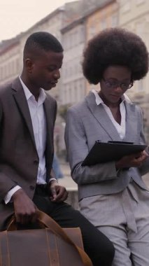 Group of two african office workers sitting on old fountain outdoors and talking about work. Attractive woman holding clipboard, handsome man with leather brown briefcase.