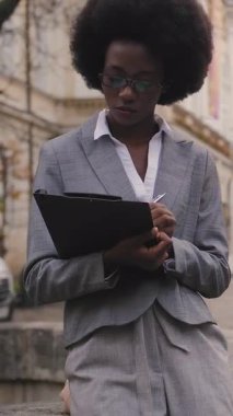 Şık takım elbiseli odaklı Afrikalı kadın şehir sokaklarında panosuyla oturuyor. Gözlüklü bir iş kadını açık havada notlar yazıyor..