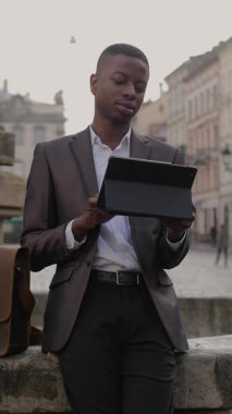 Siyah takım elbiseli genç adam şehrin eski çeşmesinde otururken dijital tablet kullanıyor. Çevrimiçi acil iş. Modern teknoloji kavramı.