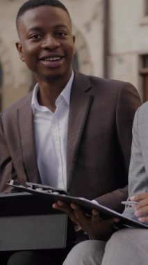 Elinde not panosu olan çekici bir kadın dijital tabletli genç bir adamın yanında ahşap bankta oturuyor. İki Afrikalı ortak dışarıda çalışma saatlerini tartışıyor..