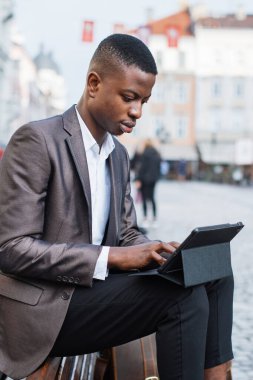 Takım elbiseli ve beyaz gömlekli genç bir siyah adam dışarıda bir bankta oturuyor ve tablet cihazına odaklanıyor.