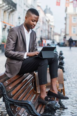 Takım elbiseli ve mokasen giyen şık bir genç adam Avrupa 'nın bir şehrinde bir bankta oturuyor, tabletine dalmış.