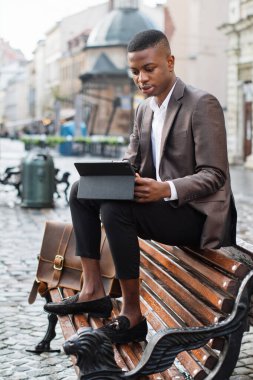 Takım elbiseli genç bir siyah adam, açık havada ahşap bir park bankında oturuyor, tabletine sarılmış, yanında deri bir çantayla.