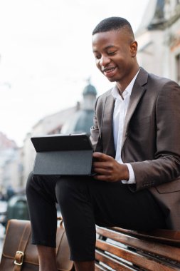 Takım elbiseli, beyaz gömlekli, gülümseyen genç bir siyah adam elinde tablet ve deri çantayla parkta bir bankta oturuyor.