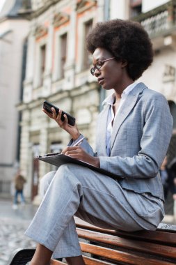 Takım elbiseli profesyonel siyah bir kadın dışarıda bir bankta oturuyor, telefonuna bakıyor ve not alıyor.