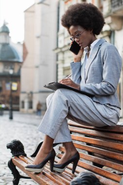 Takım elbiseli profesyonel bir siyahi kadın parktaki bankta oturur, telefonuyla konuşur ve not alır.