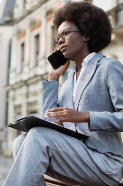 Takım elbiseli profesyonel bir siyahi kadın elinde bir dosya ve kalemle dışarıda telefonla konuşuyor.