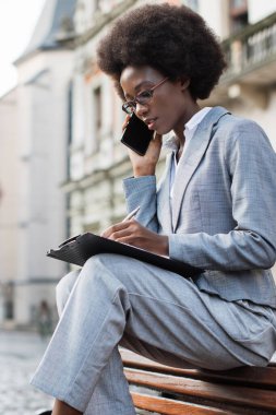 Takım elbiseli profesyonel siyah bir kadın dışarıda bankta oturuyor, akıllı telefonuyla konuşuyor ve bir panoya yazı yazıyor.
