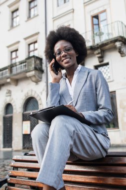 Takım elbiseli profesyonel siyah bir kadın parktaki bankta oturuyor, telefonuyla konuşuyor ve elinde kalemle bir not panosu tutuyor.
