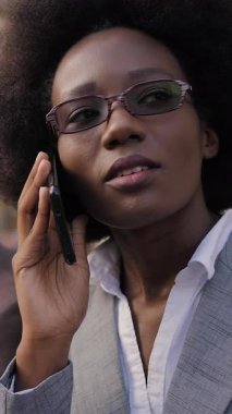 Attractive african woman in business suit sitting on wooden bench and talking on mobile. Serious lady having conversation on fresh air during break at work.