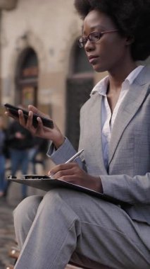 Afro-Amerikalı iş kadını modern akıllı telefonu kullanıyor ve bankta otururken panoya yazı yazıyor. Şehir caddesinde çalışan şık takım elbiseli güzel genç bir kadın..
