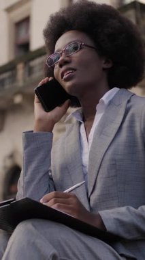 Şık giyinen, gözlüklü, akıllı telefondan konuşan ve panoya yazı yazan başarılı bir iş kadını. Afro-Amerikalı kadın bankta oturuyor ve dışarıda çalışıyor..