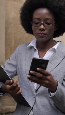 Attractive business african woman standing on city street with clipboard and modern smartphone in hands. Busy lady in suit and eyeglasses solving working issues online.