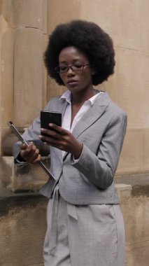 Attractive business african woman standing on city street with clipboard and modern smartphone in hands. Busy lady in suit and eyeglasses solving working issues online.
