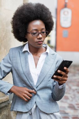 Takım elbiseli, gözlüklü profesyonel bir zenci kadın şehir caddesinde dikilirken telefonuna bakar.