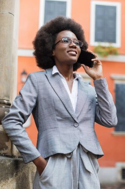 Siyah saçlı, gözlüklü ve gri takım elbiseli profesyonel bir kadın binanın önünde dikilirken akıllı telefonuyla konuşuyor.