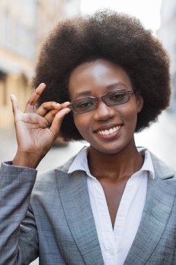 Takım elbiseli, kendine güvenen siyah bir kadın, gözlüğünü açık havada gülümserken, profesyonelliğini ve tarzını gösterirken ayarlıyor.