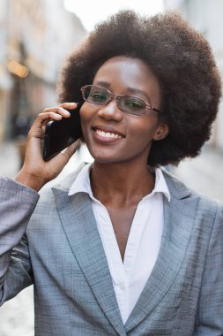 Afro saçlı ve gözlüklü profesyonel bir Afro-Amerikalı kadın şehir caddesinde akıllı telefonuyla konuşurken gülümsüyor.