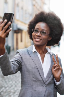Takım elbiseli profesyonel bir siyahi kadın, şehir sokaklarında barış işareti jesti yaparken telefonuyla selfie çekiyor.