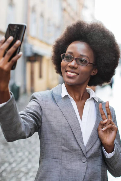 Takım elbiseli profesyonel bir siyahi kadın, şehir sokaklarında barış işareti jesti yaparken telefonuyla selfie çekiyor.