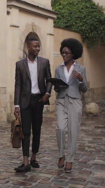 Resmi giyinmiş iki Afrikalı iş adamının uzun bir portresi. Şehir caddesinde yürüyor ve konuşuyor. Kahverengi evrak çantası taşıyan yakışıklı genç adam, elinde pano tutan kadın..
