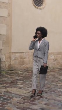 Full length portrait of stylish business lady in eyeglasses having mobile conversation while walking on city street. African woman carrying clipboard and talking outdoors.