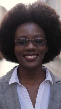 Açık havada dururken gözlükleriyle birbirine dokunan şık takım elbiseli Afro-Amerikan kadın portresi. Başarılı bir kadın gülümsüyor ve kameraya bakıyor..