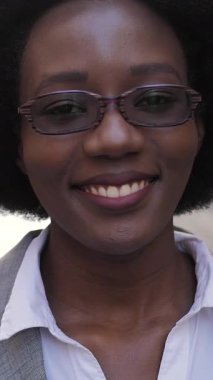 Açık havada dururken gözlükleriyle birbirine dokunan şık takım elbiseli Afro-Amerikan kadın portresi. Başarılı bir kadın gülümsüyor ve kameraya bakıyor..
