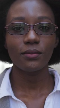 Açık havada dururken gözlükleriyle birbirine dokunan şık takım elbiseli Afro-Amerikan kadın portresi. Başarılı bir kadın gülümsüyor ve kameraya bakıyor..