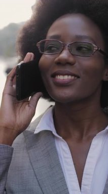 Gözlüklü ve takım elbiseli çekici afro iş kadını cep telefonuyla konuşuyor ve caddede dikilirken yana bakıyor. İnsan kavramı, iş ve sohbet.