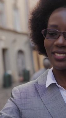 Çekici Afrikalı iş kadını modern akıllı telefondaki video görüşmesi sırasında gülümsüyor ve el kol hareketi yapıyor. Gözlüklü ve takım elbiseli mutlu kadın şehir caddesinde duruyor ve cep telefonuyla konuşuyor..