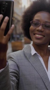 Çekici Afrikalı iş kadını modern akıllı telefondaki video görüşmesi sırasında gülümsüyor ve el kol hareketi yapıyor. Gözlüklü ve takım elbiseli mutlu kadın şehir caddesinde duruyor ve cep telefonuyla konuşuyor..