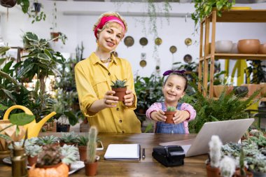 Bir kadın ve genç bir kız, parlak, davetkar bir çiçekçide saksı saksısı tutuyor.