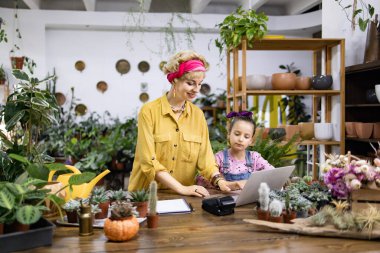 Bir kadın ve bir kız çiçekçi dükkanında birlikte çalışıyorlar. Etrafı çiçek ve bitkilerle çevrili, dizüstü bilgisayar ve not defteri kullanıyorlar.