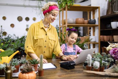 Bir anne ve kızı bir fabrikada birlikte çalışıyorlar, bir dizüstü bilgisayar kullanıyorlar ve etrafları bitki ve çiçeklerle çevrili.