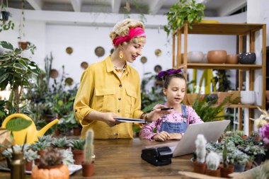 Bir kadın ve bir kız, çeşitli bitki ve saksılarla çevrili bir fabrikada dizüstü bilgisayara bakıyorlar.