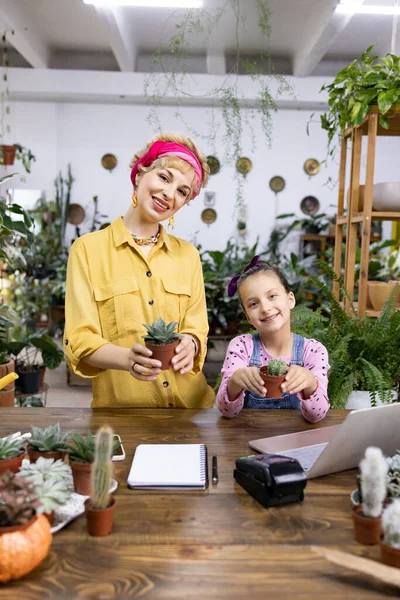Bir kadın ve genç bir kız bir bitki dükkanında saksı suyu tutarken gülümsüyorlar.