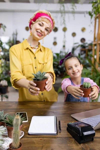 Bir kadın ve genç bir kız ellerinde küçük saksılar tutarken gülümsüyor, iç açıcı bir sahne yaratıyorlar.