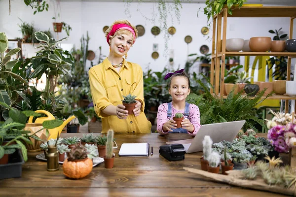 Bir kadın ve genç bir kız bir çiçekçide saksı tutarken gülümsüyorlar.