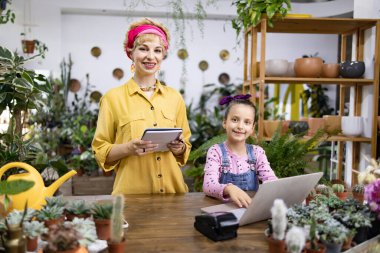 Yeşillik ve çiçeklerle çevrili bir çiçekçide çalışırken bir kadın ve bir kız gülümsüyor.