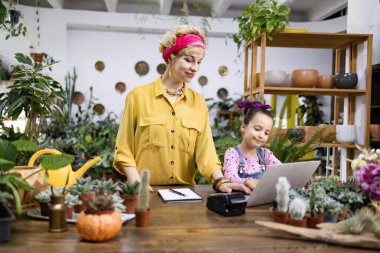 Bir kadın ve genç bir kız bir fabrikada birlikte çalışıyorlar, ahşap bir masada dizüstü bilgisayar kullanıyorlar.