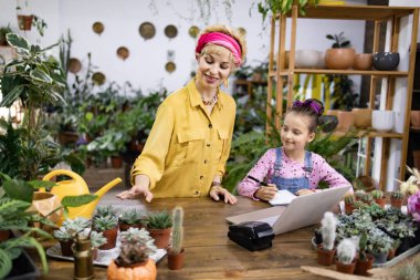 Bir kadın ve bir kız, çeşitli saksı bitkileri ve yeşilliklerle çevrili bir fabrikada birlikte çalışıyorlar.
