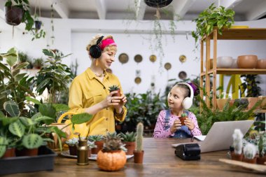 Bir kadın ve bir kız bir fabrikada, ikisi de kulaklık takıyor ve ellerinde küçük saksılar tutuyorlar.