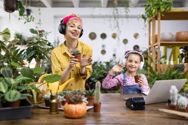 Bir kadın ve bir kız, kulaklık takıp kameraya gülümserken çiçek tutuyorlar.