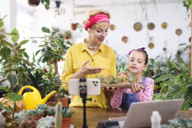 Bir kadın ve bir kız bitkilerle dolu, parlak bir odada hava santralleriyle ilgili bir video çekiyorlar.